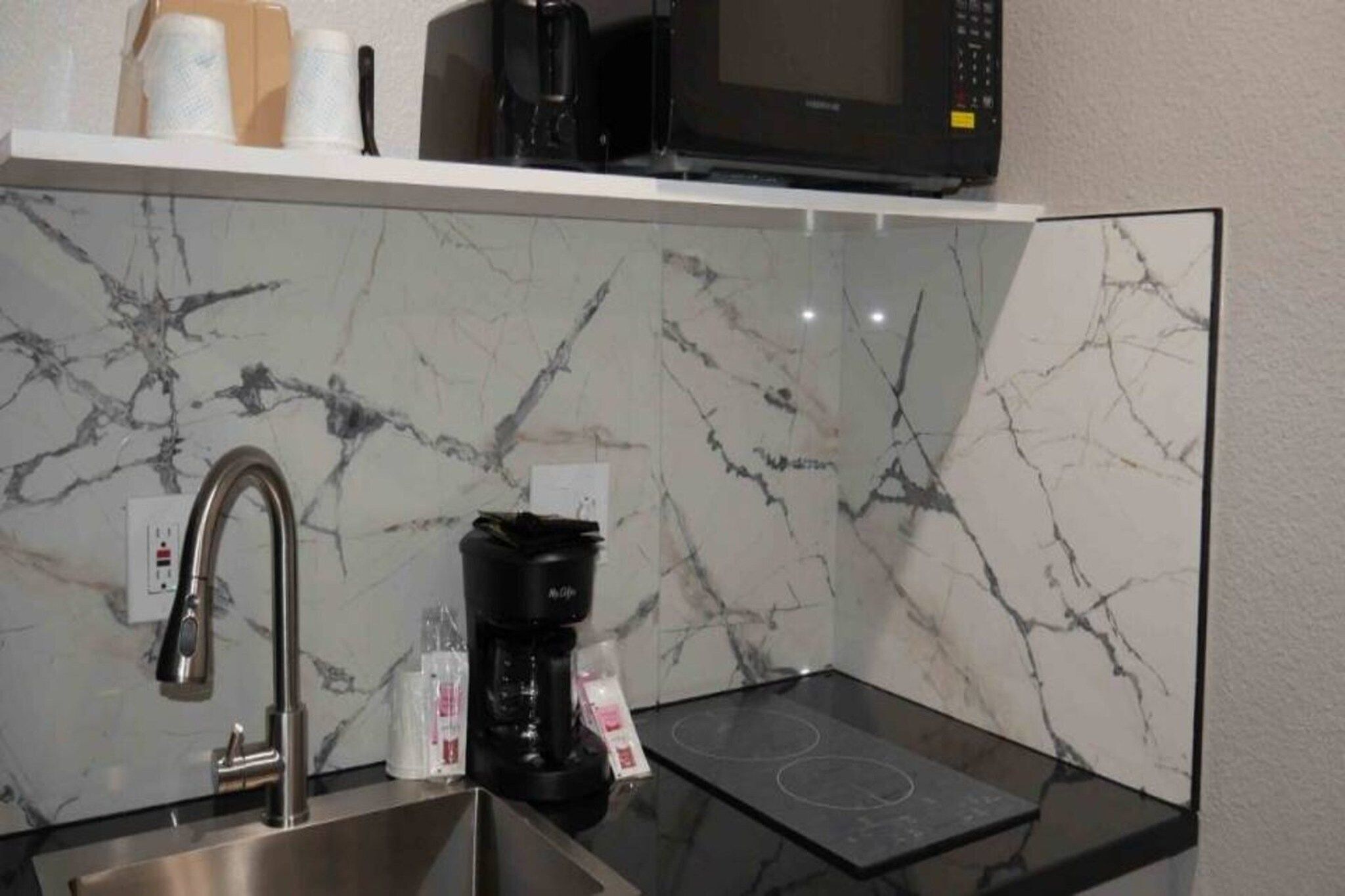 Una cocina con un lavamanos de acero inoxidable, una cafetera negra, un microondas y un protector contra salpicaduras de mármol.