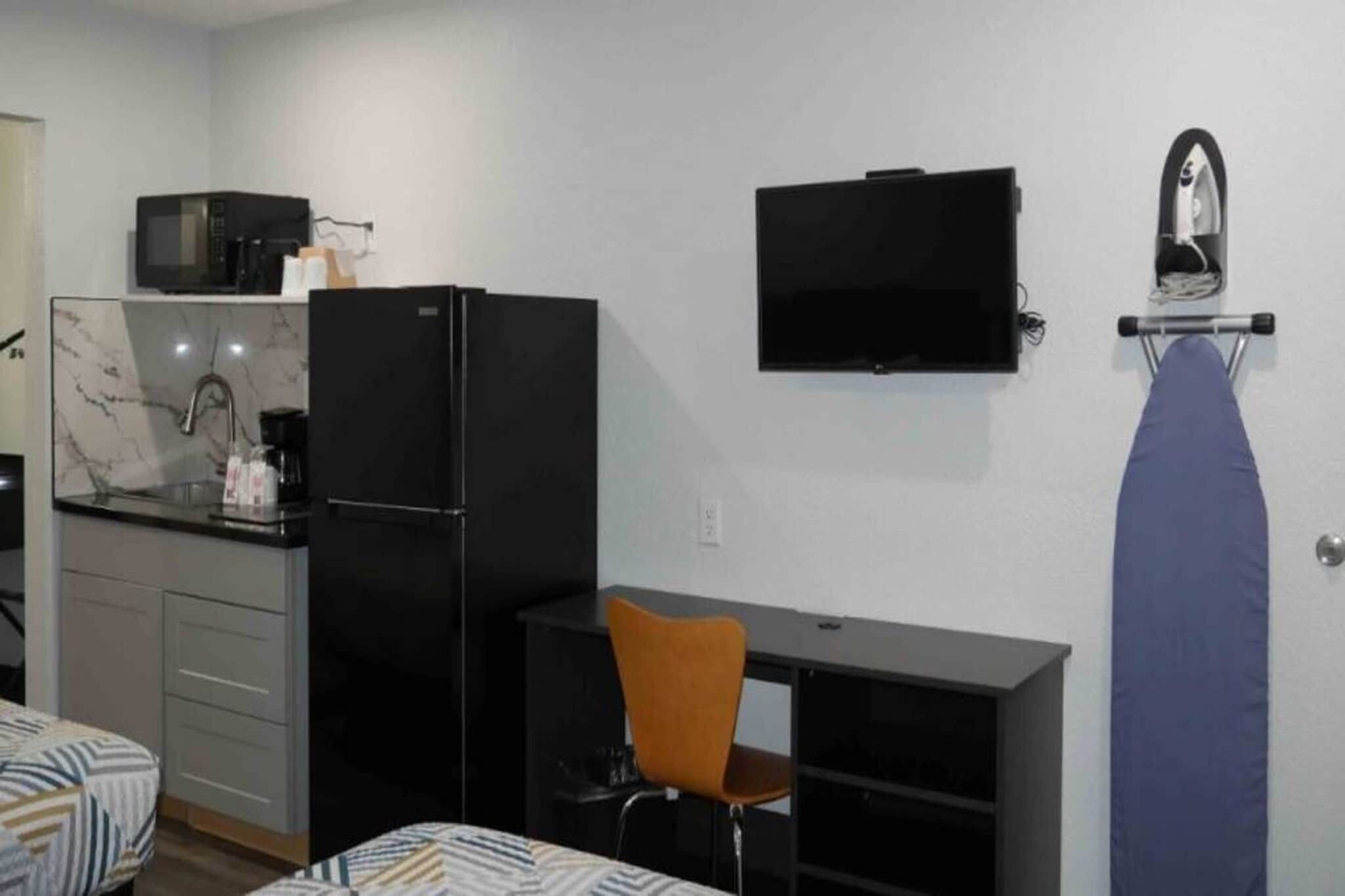 A compact hotel room with a kitchenette, a bed with patterned bedding, a desk with a chair, and a TV mounted on the wall.