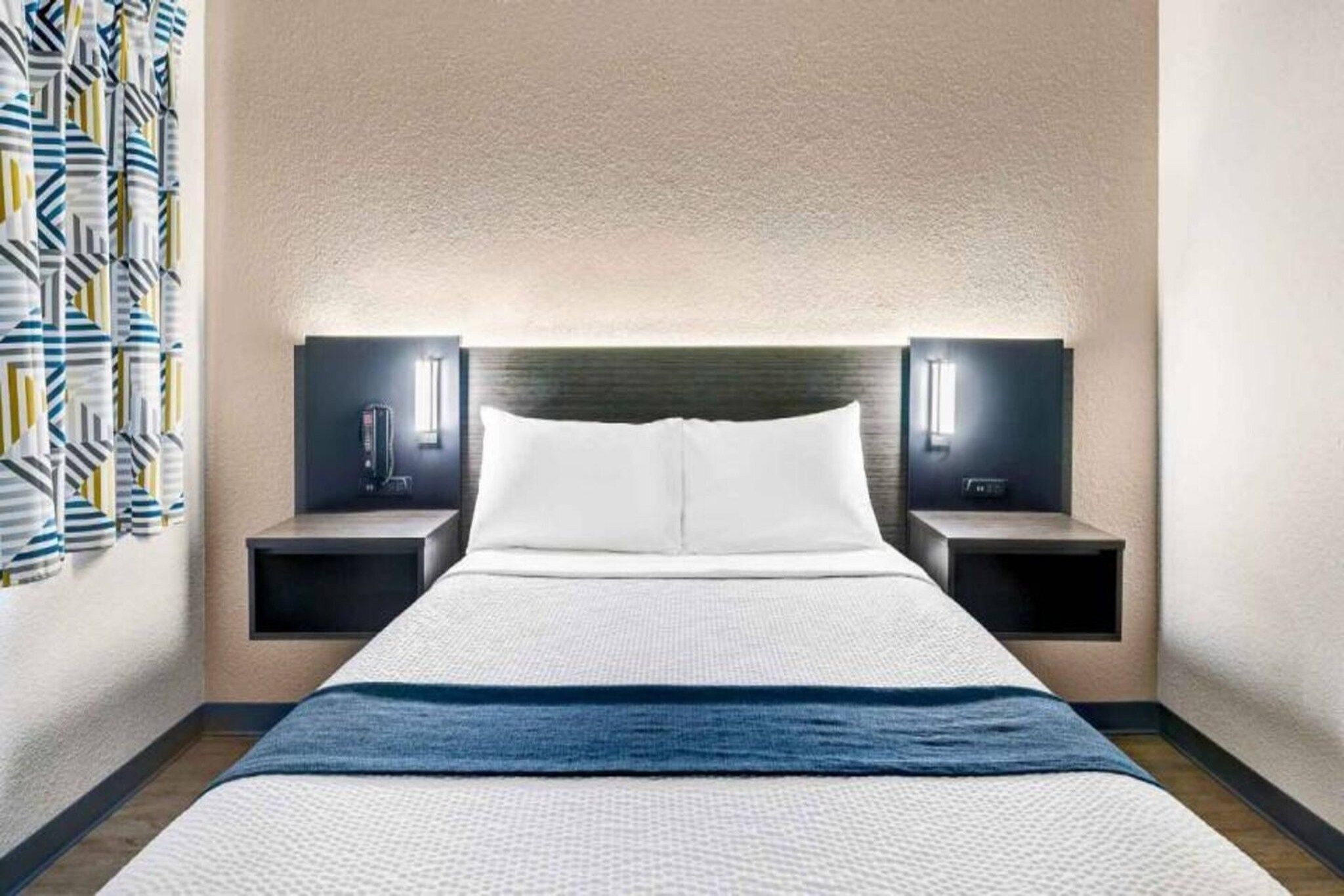 Hotellihuone, jossa on sänky, kaksi yöpöytää, sängynpääty ja seinään kiinnitetty valaisin.