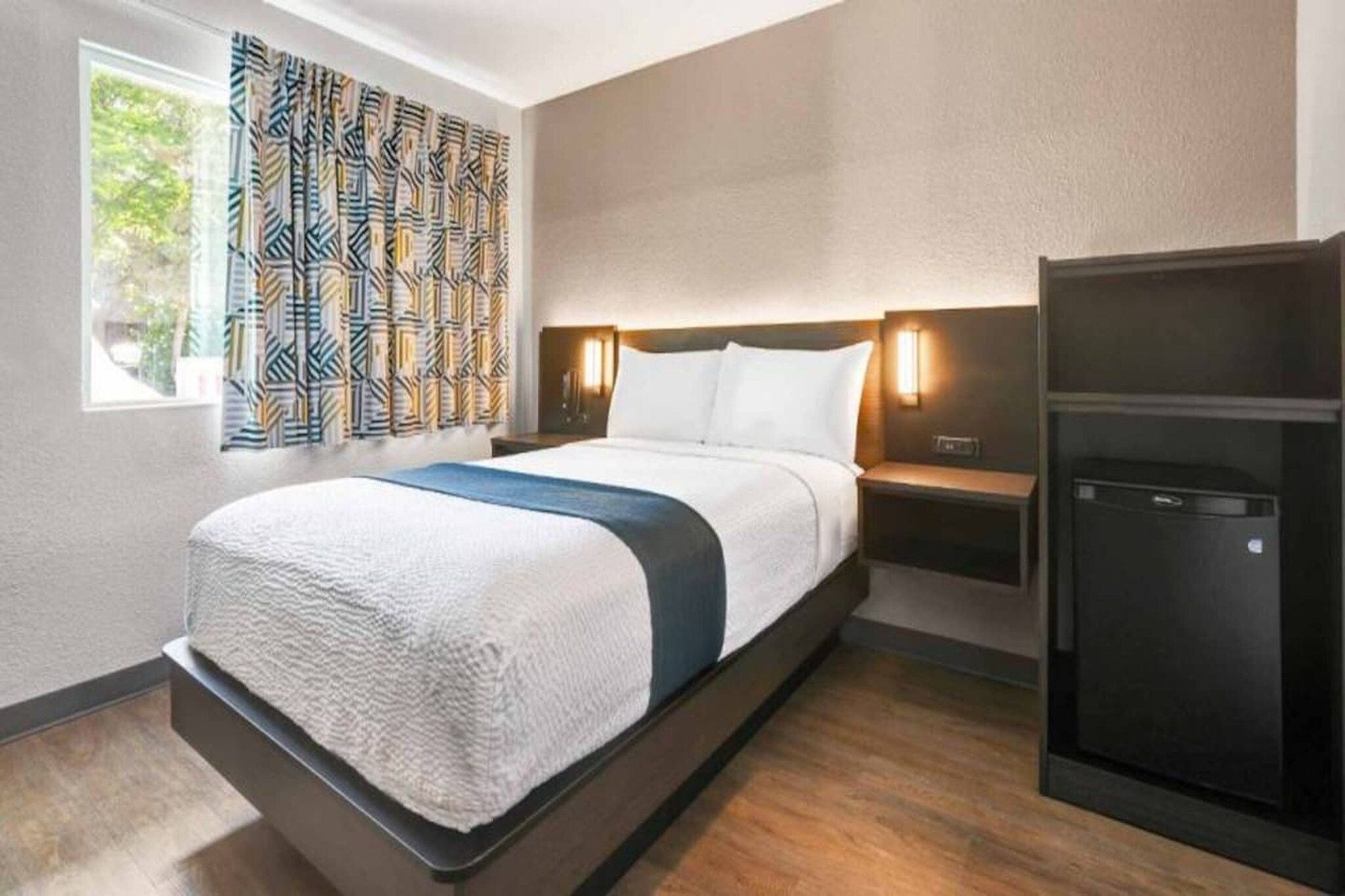 Una habitación de hotel con una cama, mesitas de noche, un armario y una ventana con cortinas estampadas.