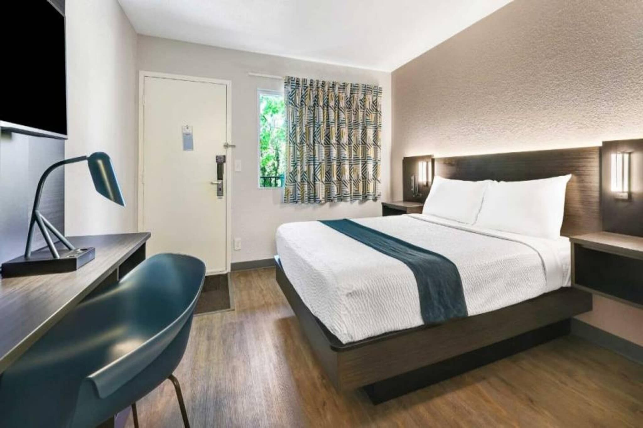 Hotellihuone, jossa on sänky, yöpöydät, työpöytä tuolilla sekä ikkuna, jossa on verhot.