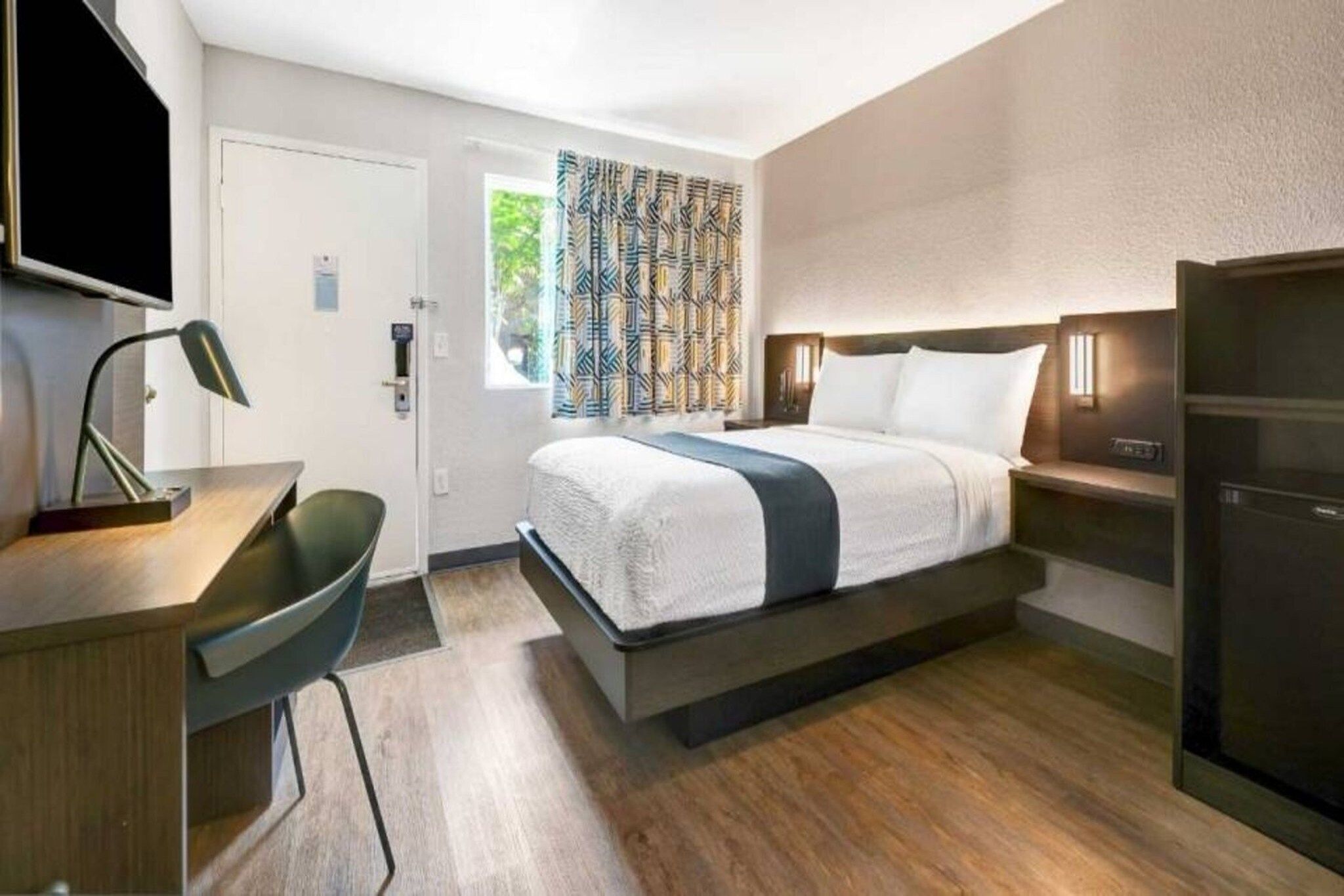 A modern hotel room with a bed, desk, chair, and a television.
