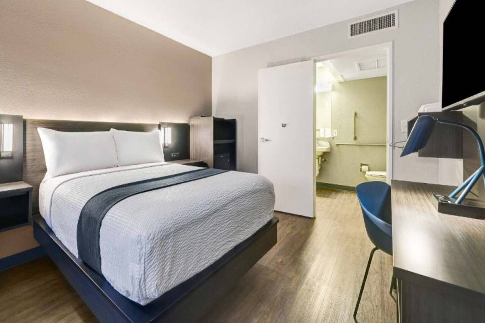 Moderni hotellihuone, jossa on suuri sänky, yöpöydät, työpöytä ja kylpyhuone, joka näkyy avoimesta ovesta.
