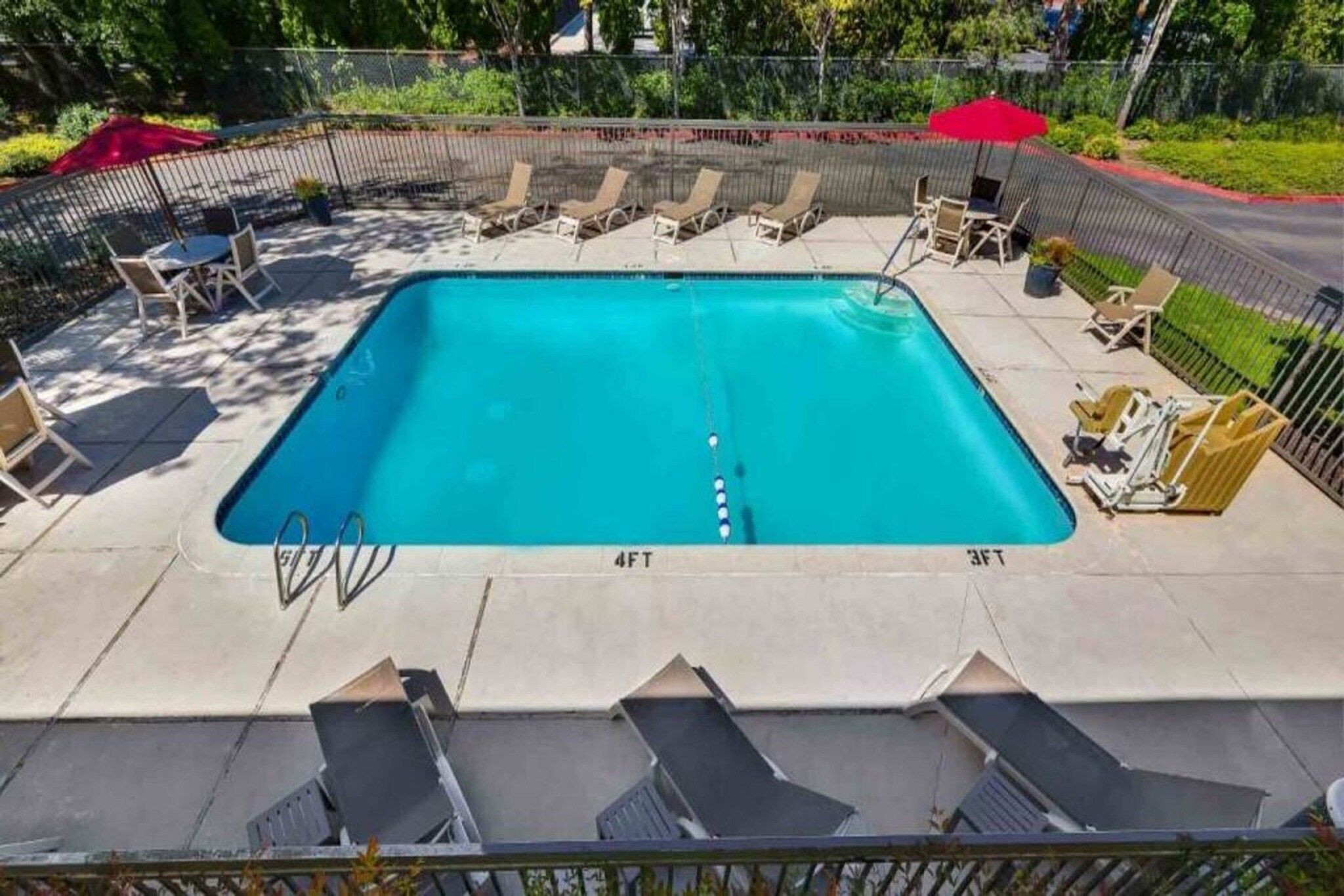 En rektangulär swimmingpool med djupmarkeringar som visar 1,2 meter och 90 centimeter, omgiven av solstolar och parasoller.
