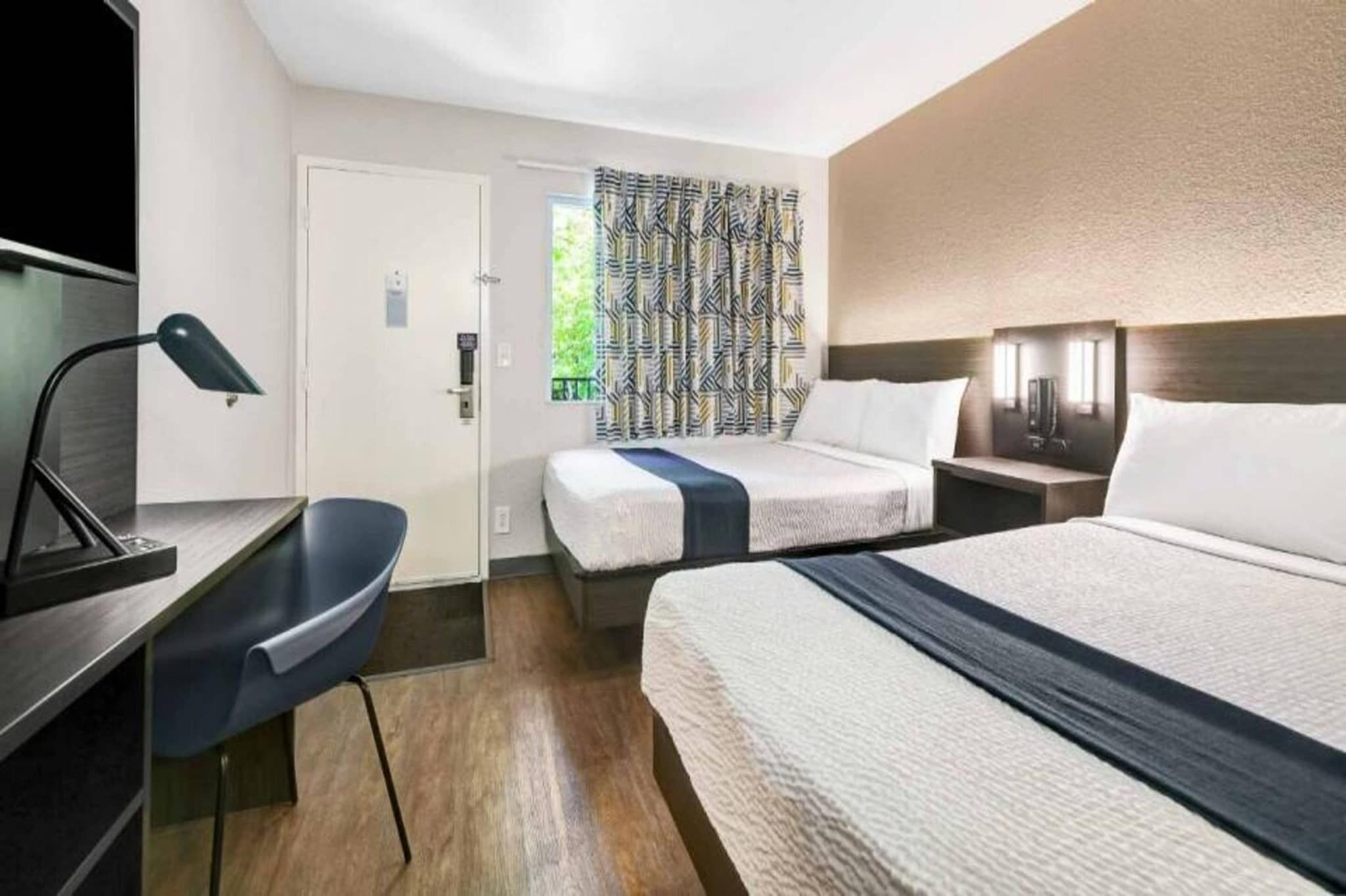 Ett hotellrum med en säng, ett sängbord, en lampa, en stol och en mönstrad gardin.