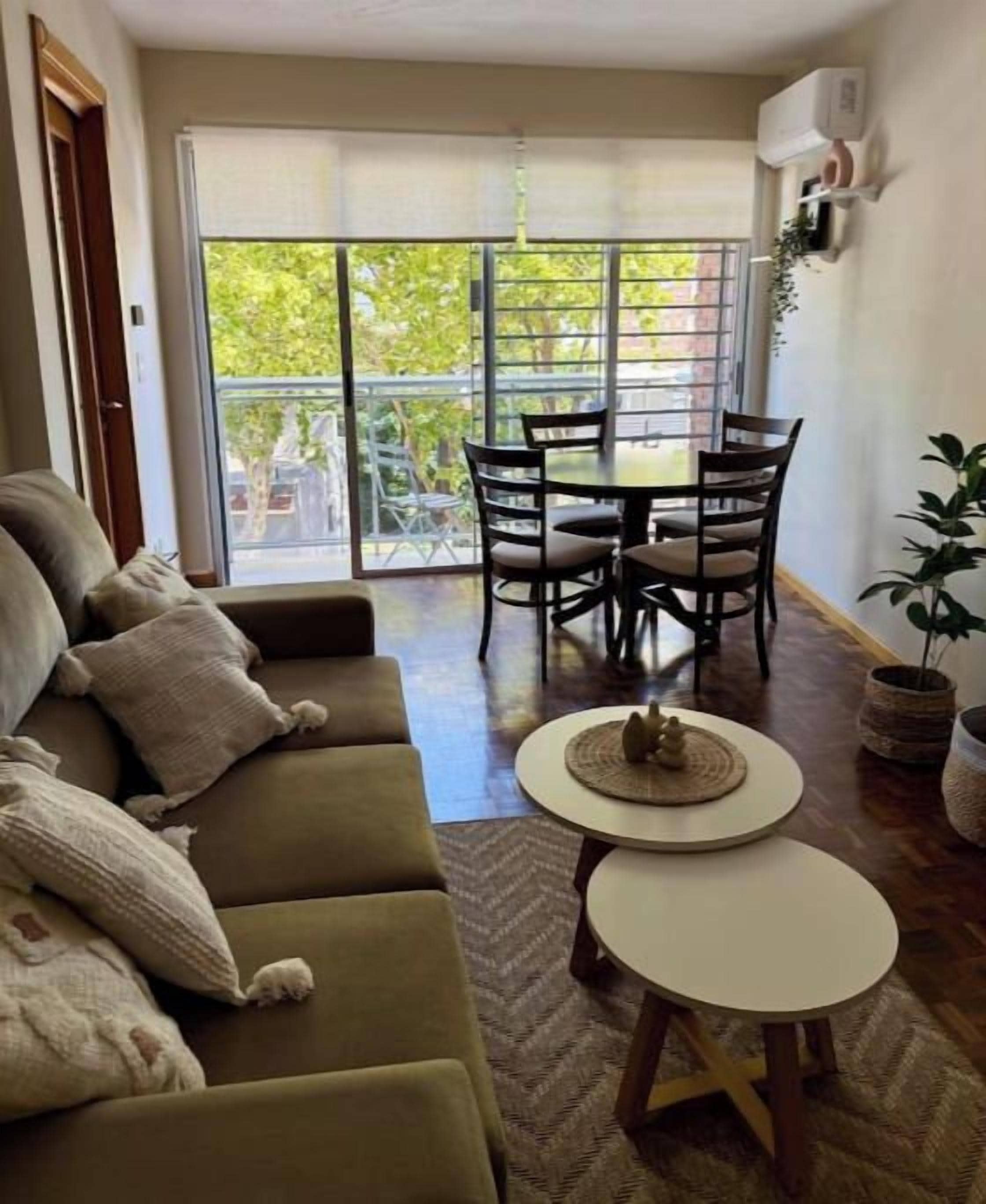 A living room with a sofa, a dining table with chairs, a small round table with a decorative item, and a sliding glass door leading to an outdoor area.