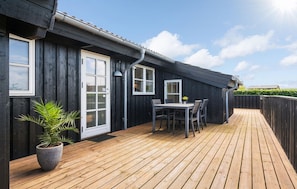 Eine Holzterrasse mit Tisch und Stühlen, einem Blumentopf und einem dunklen Gebäude mit weiß gefassten Fenstern.