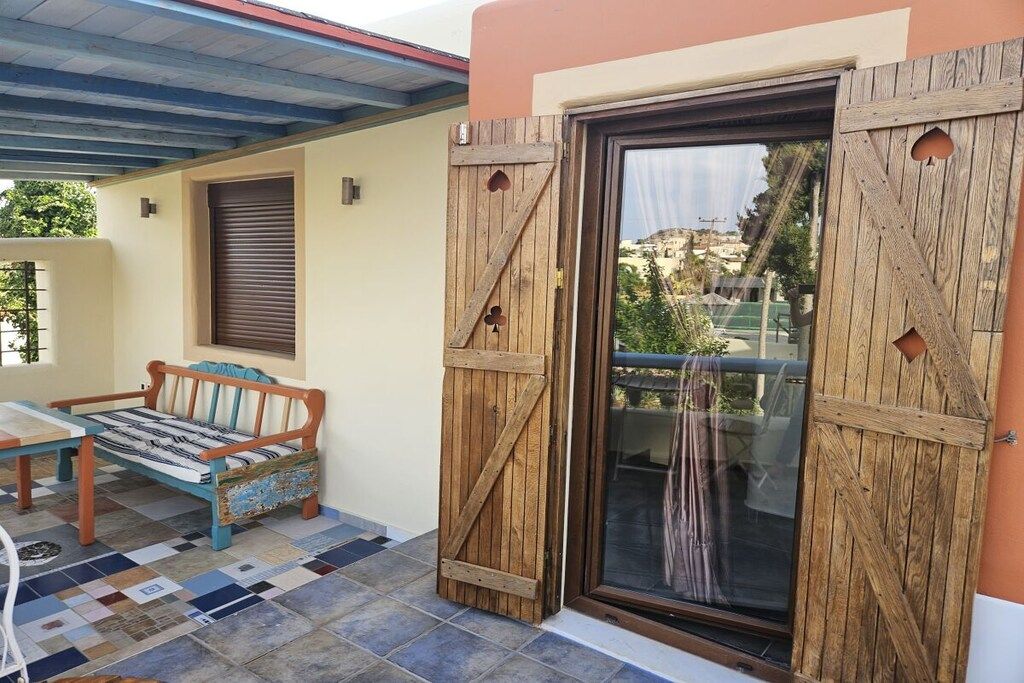 Un espace extérieur abrité avec des sièges, comprenant un banc en bois et une table, une porte-fenêtre coulissante avec un cadre en bois et un sol carrelé à motifs.
