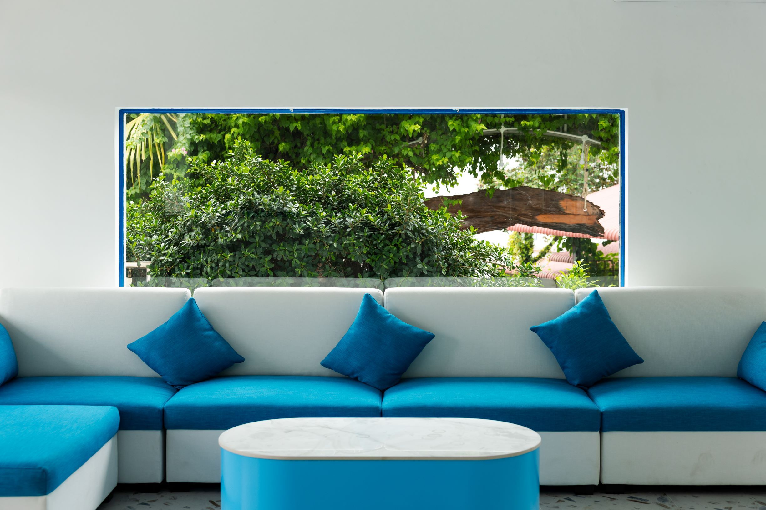 Un salón moderno con un sofá blanco y cojines azules, una mesa de centro redonda y un ventanal con vistas a follaje verde.