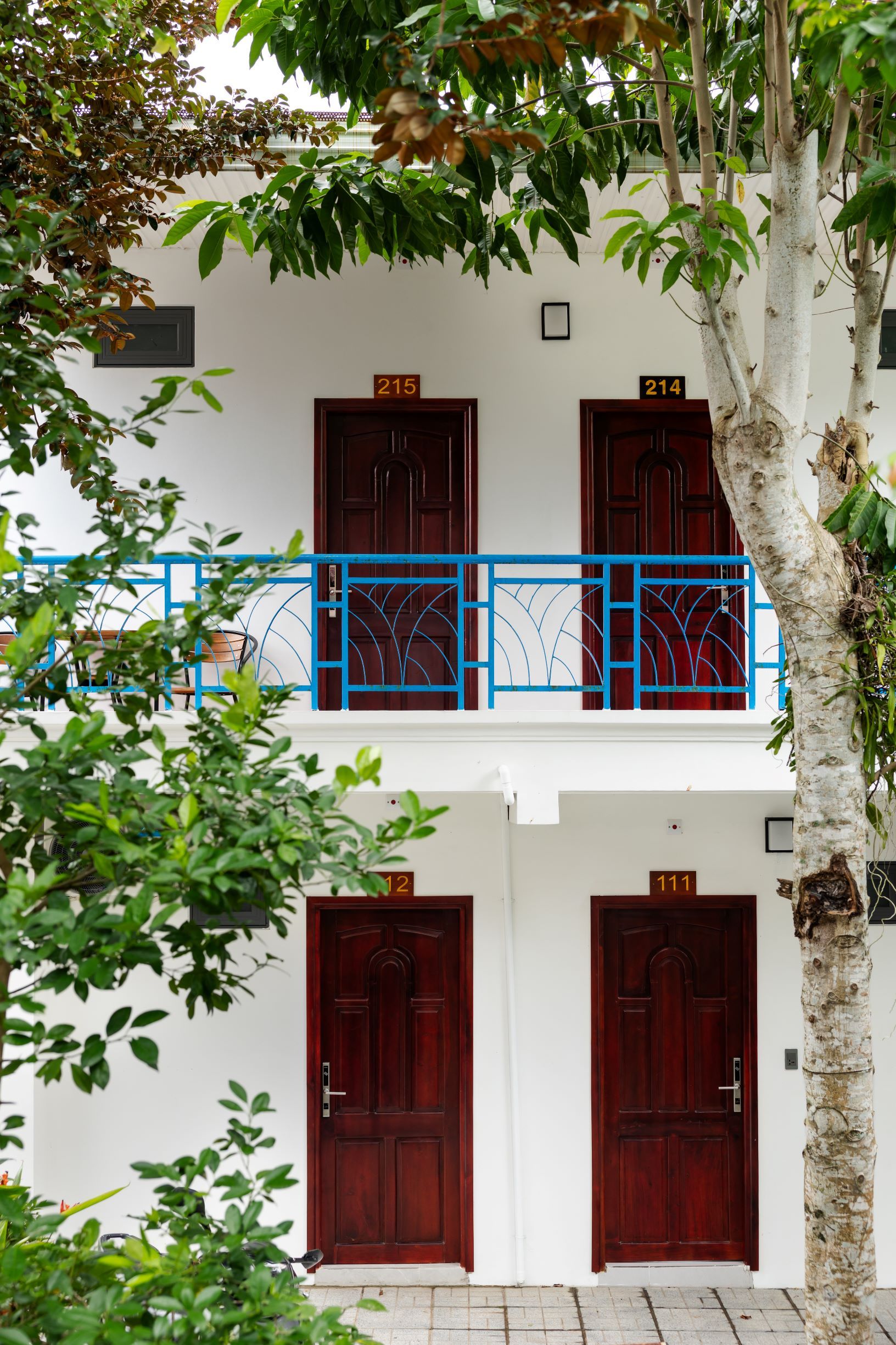 A two-story building with numbered doors, a blue balcony railing, and a tree in the foreground.