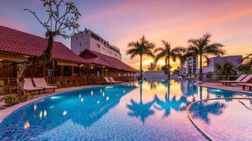 A swimming pool with lounge chairs, palm trees, and buildings with red-tiled roofs.
