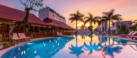 A swimming pool with lounge chairs, palm trees, and buildings with red-tiled roofs.