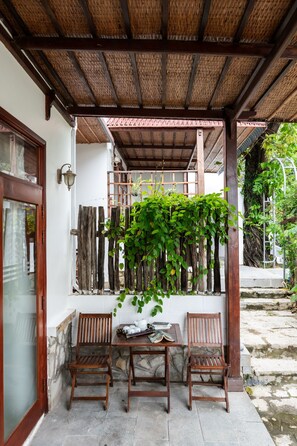 Un espacio de estar al aire libre cubierto con dos sillas de madera y una mesa, techo de bambú y una pared con plantas trepadoras.