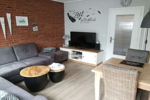A modern living room with a grey sofa, a wooden coffee table, a TV, and a dining area with a wicker chair. - Holiday apartment with 1 bedroom (Westerland)