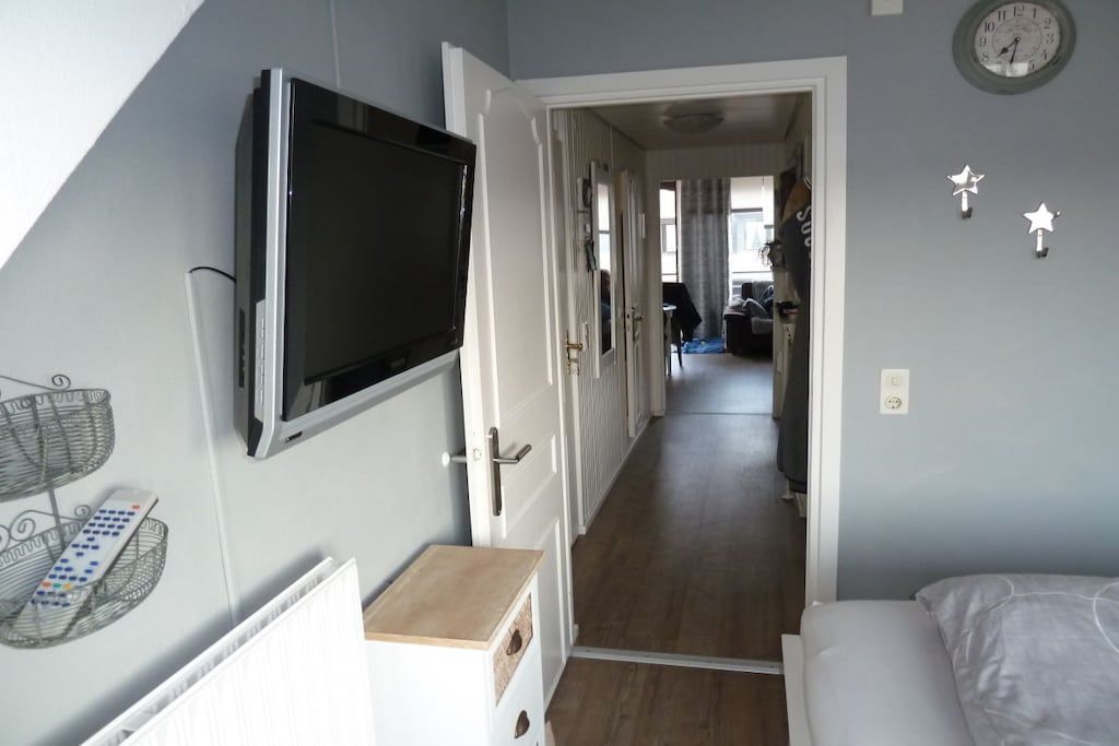 A narrow hallway with a television mounted on the wall, a door leading to another room, and a bed at the end.