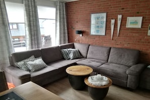 A living room with a grey sectional sofa, two round coffee tables, a brick wall, and a window with curtains. - Holiday apartment with 1 bedroom (Westerland)