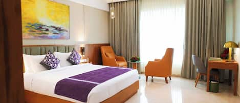 A hotel room with a large bed, a desk, two chairs, a small table with flowers, and a painting on the wall.