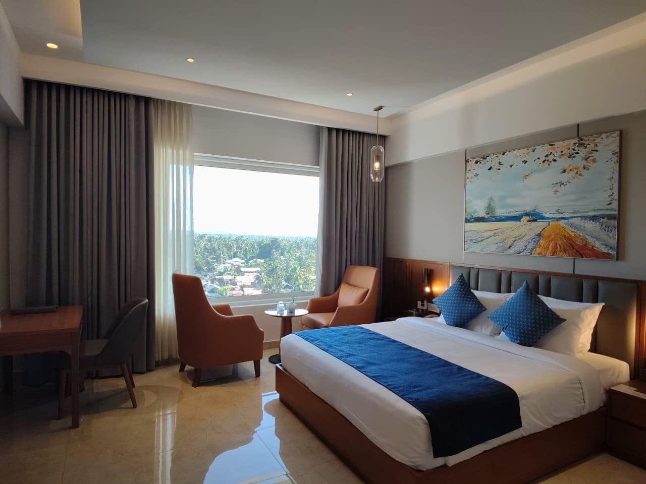Quarto de hotel com uma cama grande, uma escrivaninha, duas cadeiras, uma mesa pequena e um quadro na parede.