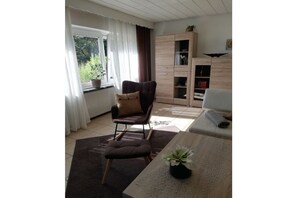 Een woonkamer met een donkerblauwe fauteuil, een houten kast en een raam met gordijnen.