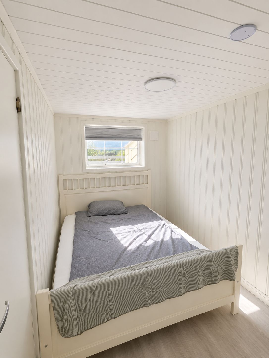 Une chambre avec un lit simple, un plafond en pente, une fenêtre avec des stores et un luminaire au plafond.