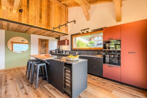 A modern kitchen with a central island, bar stools, and a view of a house through a window. - Chalet Haute Cime - Les Gets - Les Cornuts (Les Gets)