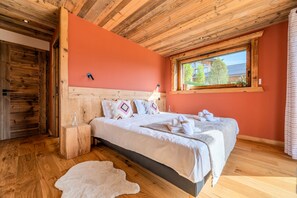 Une chambre avec un plafond en bois, un grand lit avec une literie blanche, une table de chevet en bois, une bouteille en verre et une vue sur la verdure à l’extérieur.