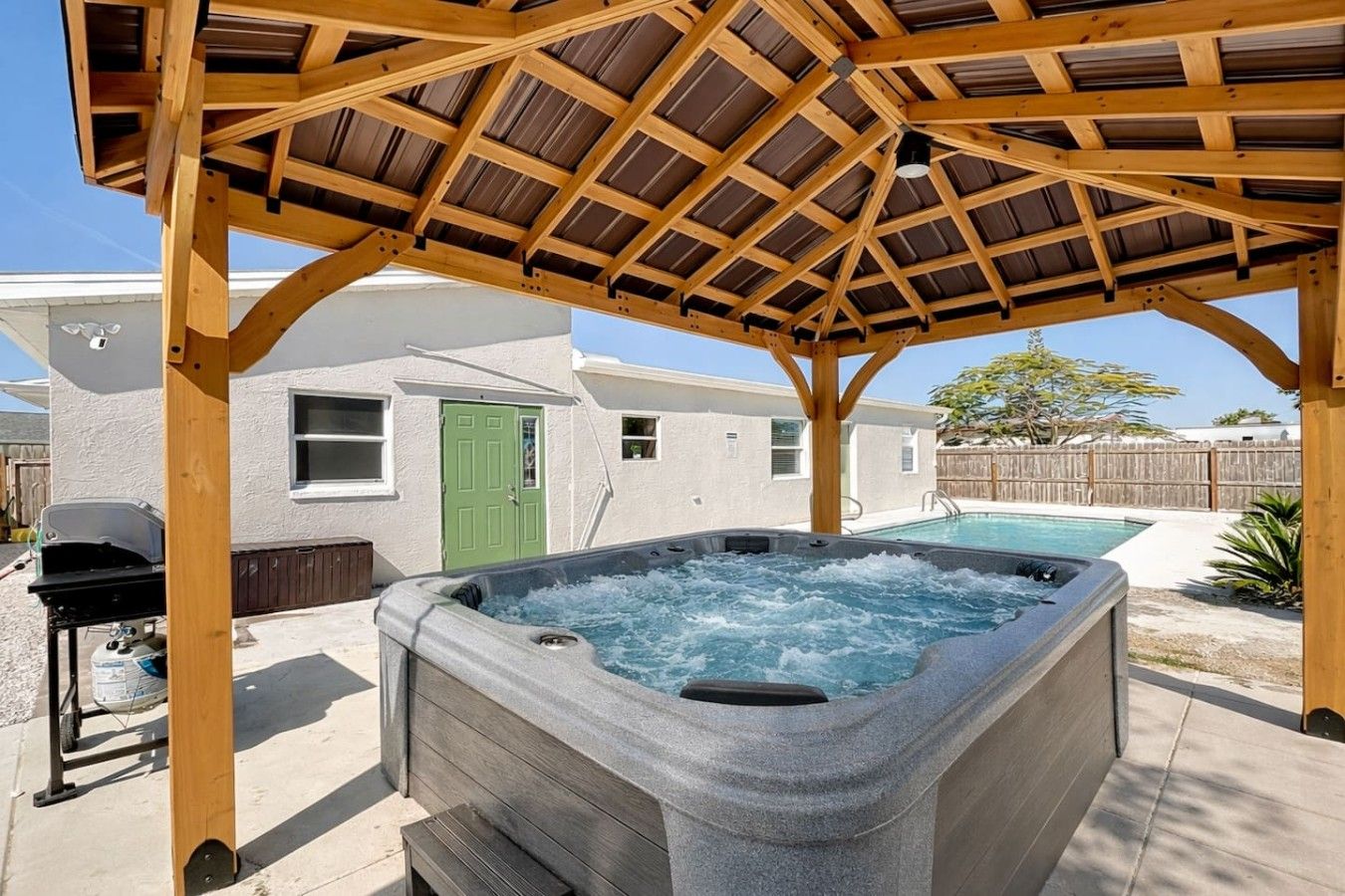 Ein Whirlpool unter einer Holzpergola in einem Garten mit Pool und einem Haus im Hintergrund.