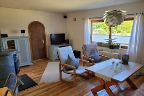 A cozy room with a wooden floor, a television, a dining table with chairs, a fireplace, and a window with a view of greenery. - Holiday home on the Baltic Sea (Glücksburg)