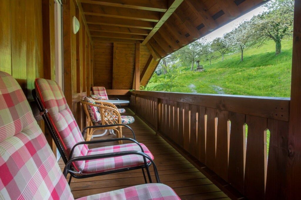 A covered porch with wooden railings, a row of checkered chairs, and a view of a green landscape with trees.