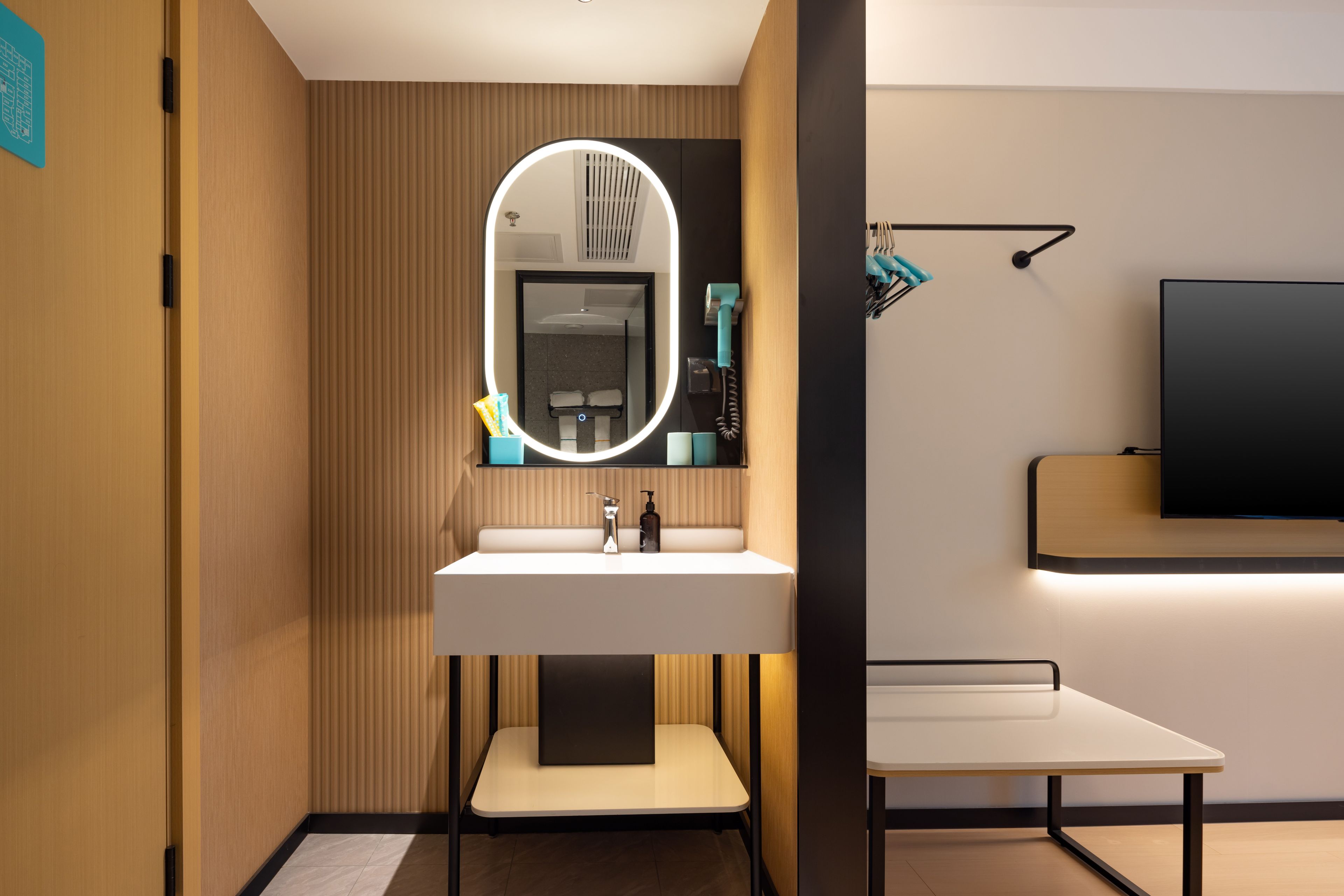 A modern bathroom with a vanity, a mirror with lighting, a towel rack, and a flat-screen TV mounted on the wall.