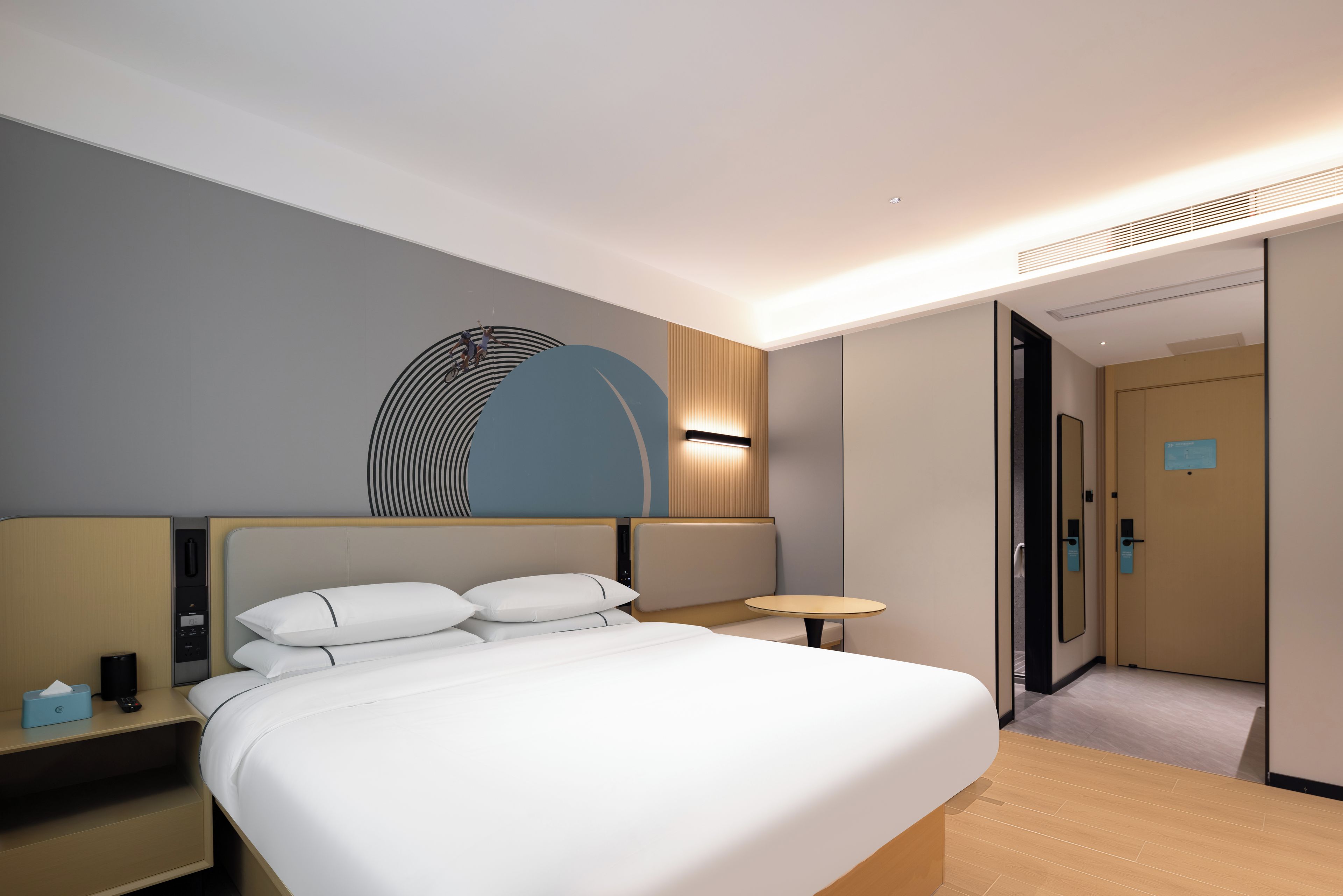A modern hotel room with a large bed, bedside tables, a wall-mounted lamp, and a door leading to another room.