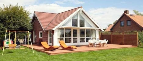 Une maison avec une grande façade vitrée, un patio avec des chaises et un portique avec des balançoires.