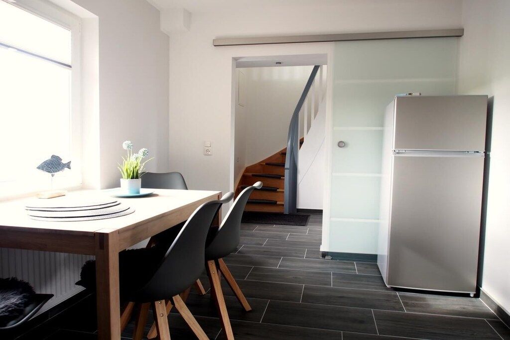 A modern kitchen with a wooden table, black chairs, a white refrigerator, and a staircase leading to another level.