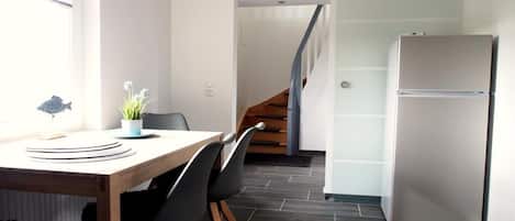 Una cocina moderna con una mesa de madera, sillas negras, un refrigerador blanco y una escalera que sube a otro nivel.