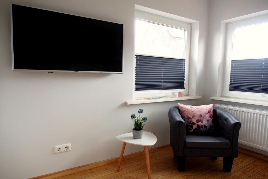 Ein Wohnzimmer mit einem an der Wand befestigten Flachbildfernseher, einem schwarzen Ledersofa mit Kissen im geometrischen Muster, einem kleinen weißen Tisch mit Topfpflanze und zwei Fenstern mit Jalousien.
