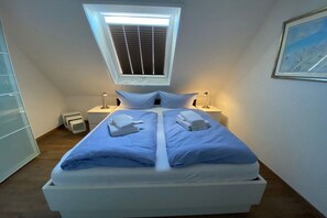 A bedroom with a skylight, a bed with blue bedding, two bedside tables with lamps, and a framed picture on the wall. - Holiday apartment with 1 bedroom (Krummhörn \/ OT Greetsiel)
