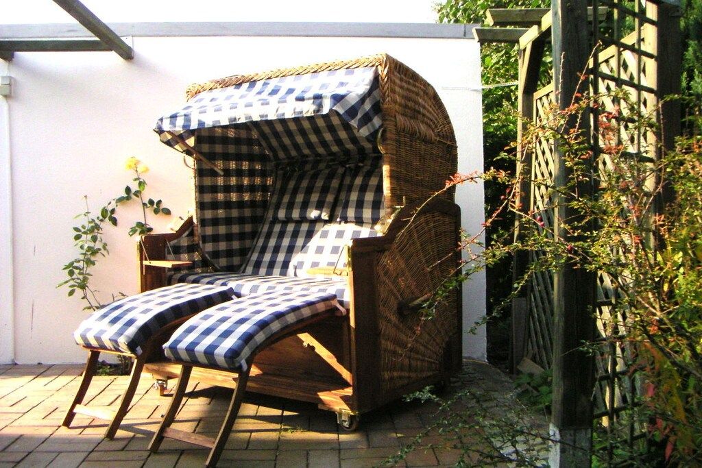 Une chaise en osier recouverte d’un tissu à carreaux, agrémentée de coussins assortis, placée à l’extérieur, avec un mur blanc et de la verdure en arrière-plan.