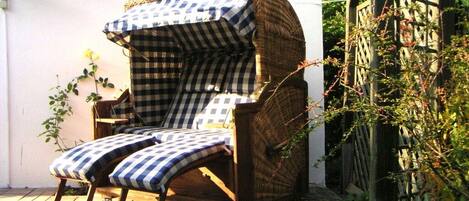 Una silla de mimbre con funda y cojines a cuadros, ubicada al aire libre frente a una pared blanca y follaje.