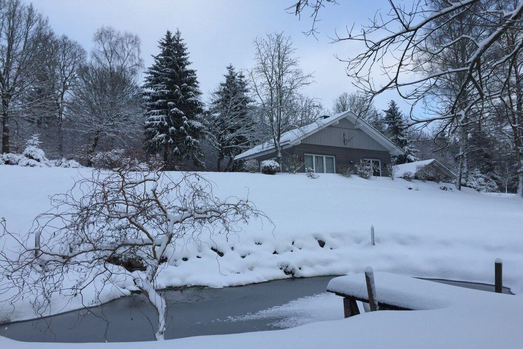 A snow-covered landscape with a house, trees, and a small body of water.