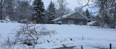 A snow-covered landscape with a house, trees, and a small body of water.