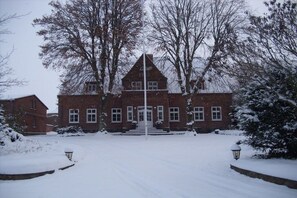 A snow-covered building with a symmetrical facade, a central entrance with steps, and a flagpole in front. - Marquardt Country House (Fehmarn)