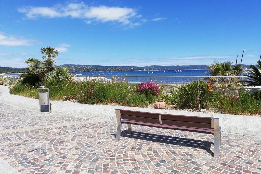 A paved walkway with a bench, leading to a beach with a clear view of the sea and distant hills.