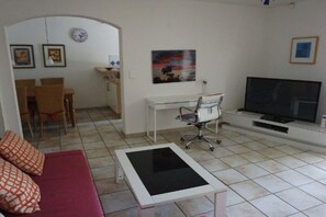 Ein Wohnzimmer mit einer Couch, einem Couchtisch, einem Schreibtisch, einem Sessel, einem Fernseher und einem Bild an der Wand.