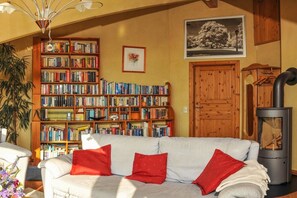 Een gezellige woonkamer met een witte bank, rode kussens, een grote boekenkast, een lijst met een afbeelding, een houten deur en een houtkachel.