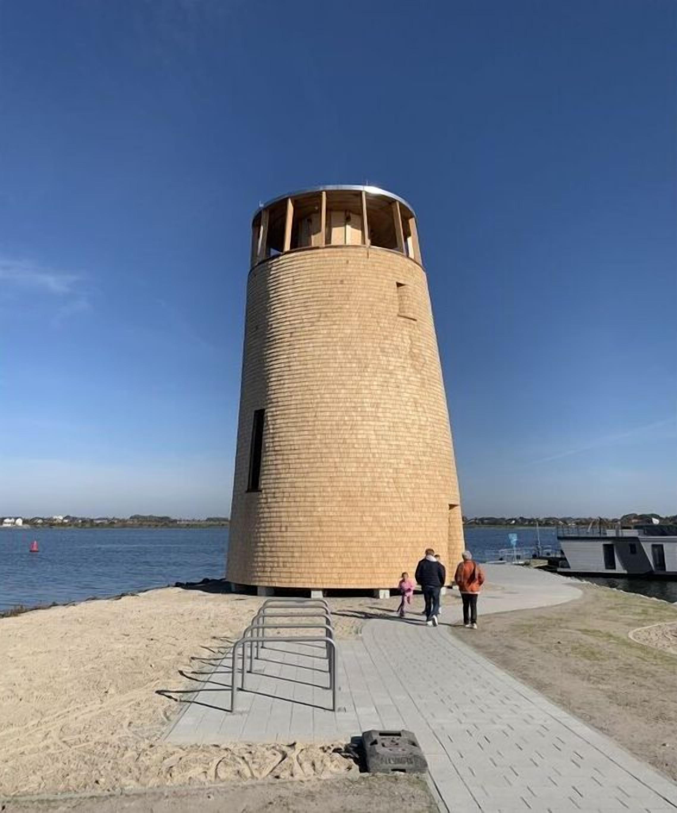 Una torre cilíndrica de ladrillo con techo plano, ubicada cerca de un muelle con un paseo pavimentado.
