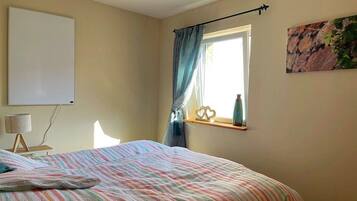 Een slaapkamer met een bed, een bedlampje, een raam met gordijnen en een aan de muur hangend schilderij.