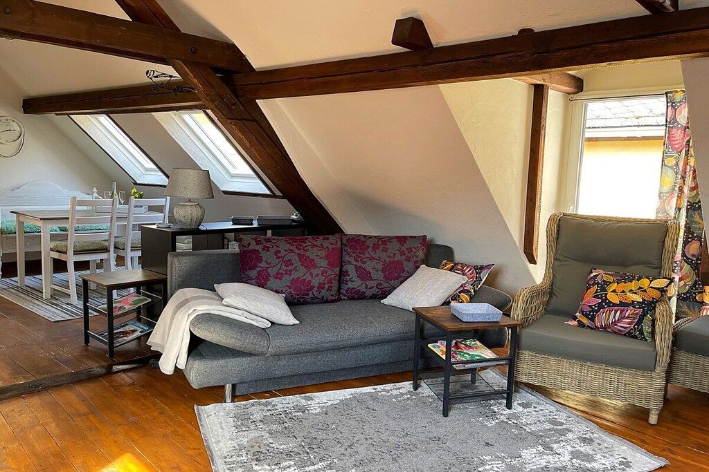Een loft-achtige kamer met een schuin dak, een grijze bank, een rieten fauteuil en een kleine tafel.
