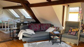 Een loft-achtige kamer met een schuin dak, een grijze bank, een rieten fauteuil en een kleine tafel.