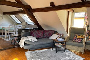 Een loft-achtige kamer met een schuin dak, een grijze bank, een rieten fauteuil en een kleine tafel.