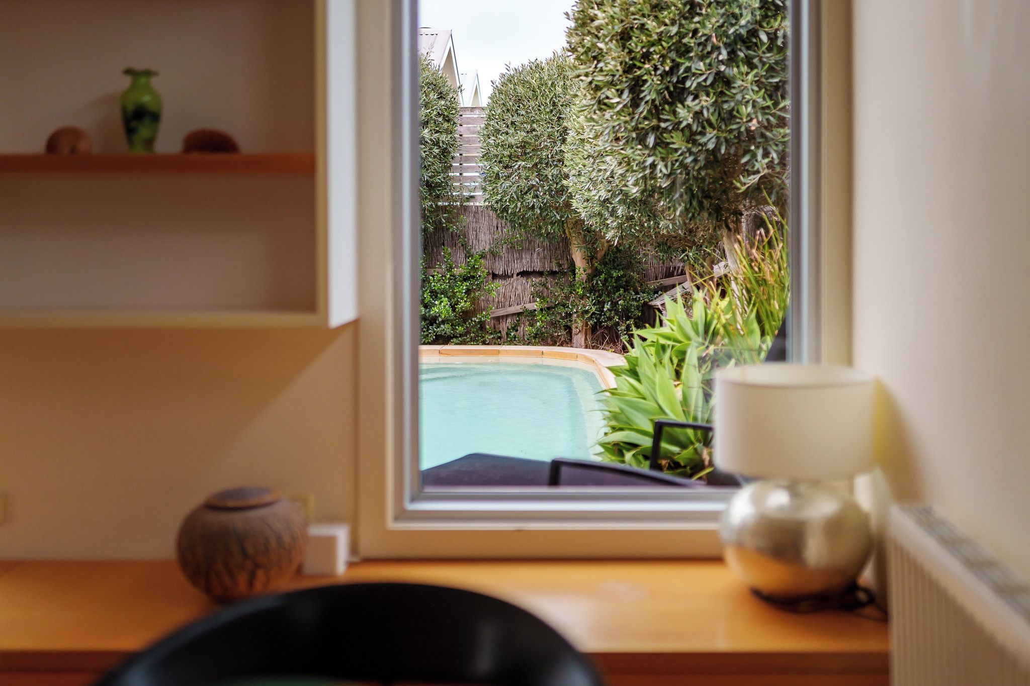 A view through a window of a pool and greenery, a wooden table with a vase and a cup, and a shelf with decorative items.